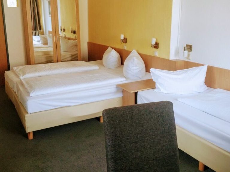 Doppelbett und ein Einzelbett im hellen Hotelzimmer mit Spiegel und Stuhl.