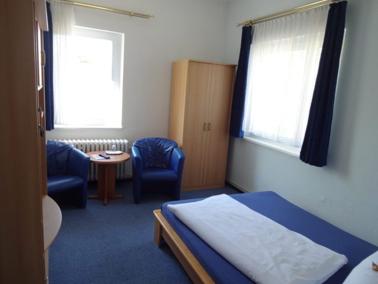 Ein einfaches Zimmer mit zwei blauen Stühlen, einem Tisch und einem Bett.
