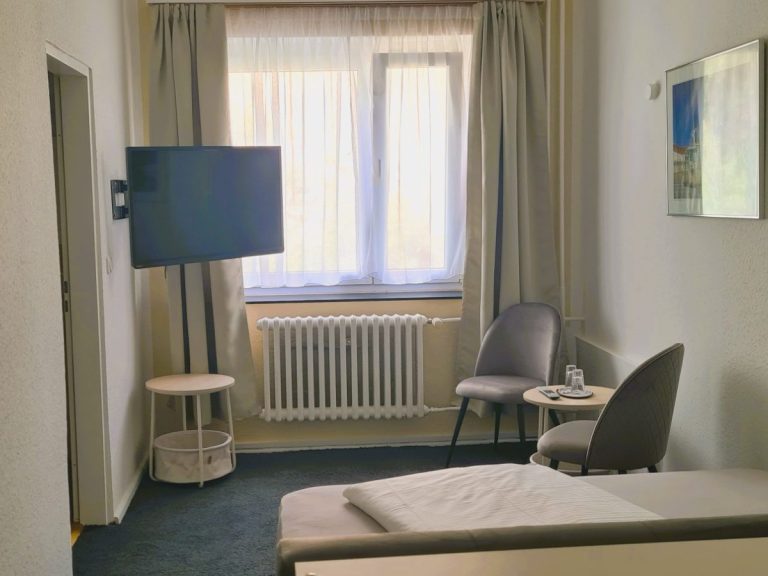Ein Bett mit grauem Kopfteil, Kopfkissen und einer weißen Decke auf einem hellen Bettbezug.