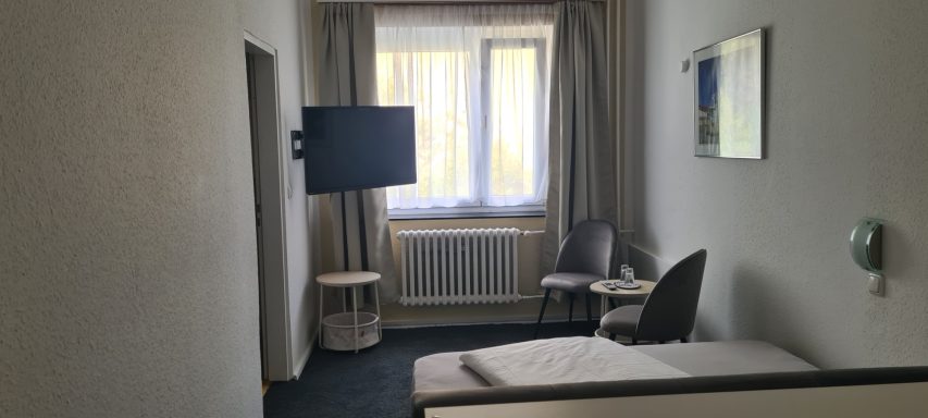 Helles Zimmer mit einem Bett, Fernseher, Schreibtisch und großen Fenstern.