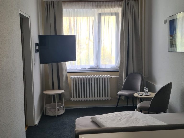 Helles Hotelzimmer mit Fernseher, Tisch, Stühlen und Fenster mit Vorhängen.