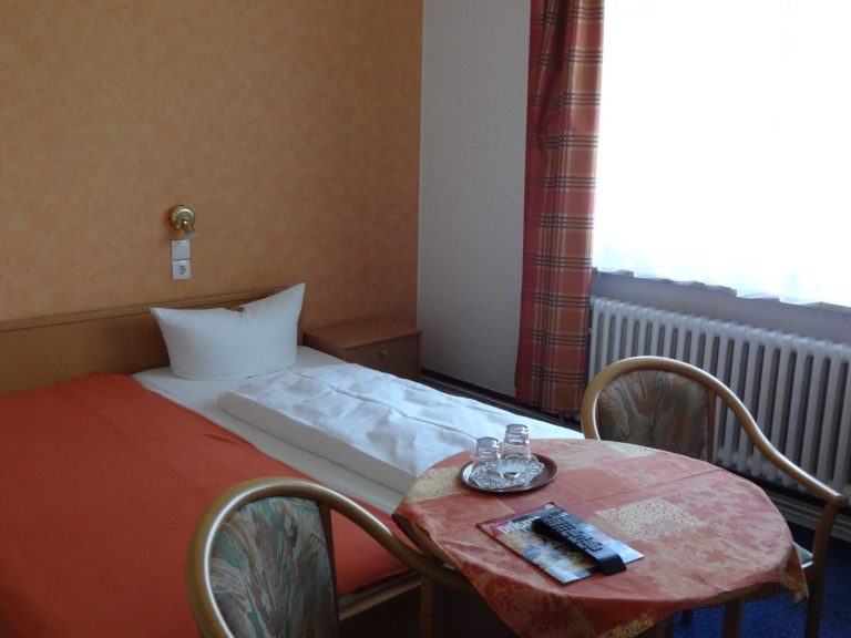 Beispiel Standard Einzelzimmer Ein einfaches Zimmer mit einem Bett, Tisch und Stühlen, sowie einem Fenster mit Vorhang.