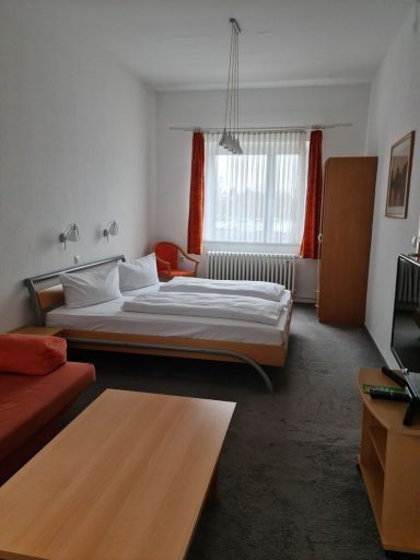 Helles Zimmer mit Doppelbett, Sofa, Tisch und TV, orangefarbene Vorhänge.