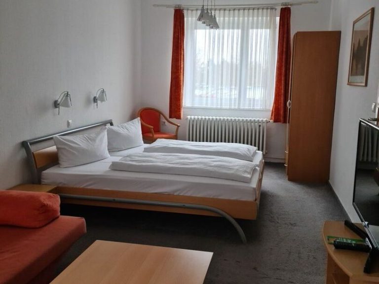 Beispiel Komfort Doppelzimmer Helles Zimmer mit einem Doppelbett, orangefarbenem Sofa, einem Fernseher und Fenster mit Vorhängen.