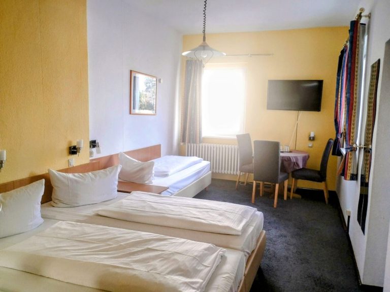 Helles Hotelzimmer mit drei Betten, Tisch, Stühlen und Fernseher.